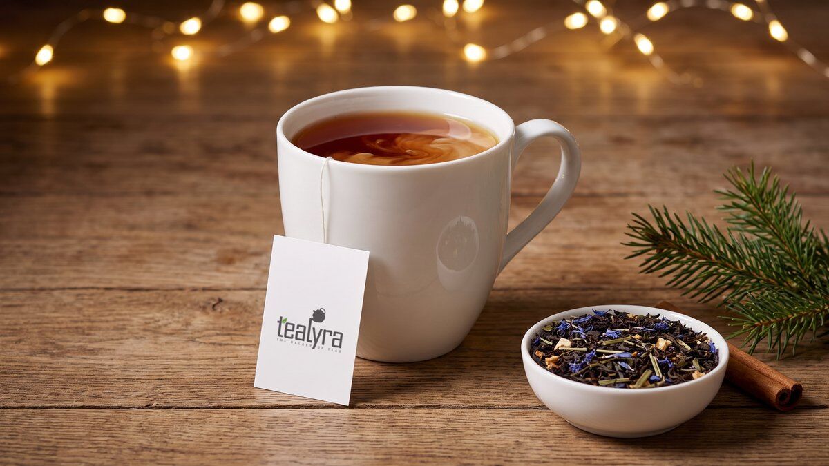 A white mug of black tea with milk and a Tealyra tea tag next to a bowl of loose Russian Earl Grey, a cinnamon stick and a fir sprig