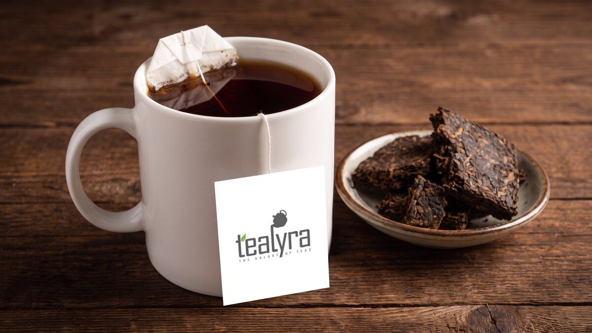 White mug of dark ripe pu-erh tea with a Tealyra tea bag tag beside broken chunks of a pu-erh brick
