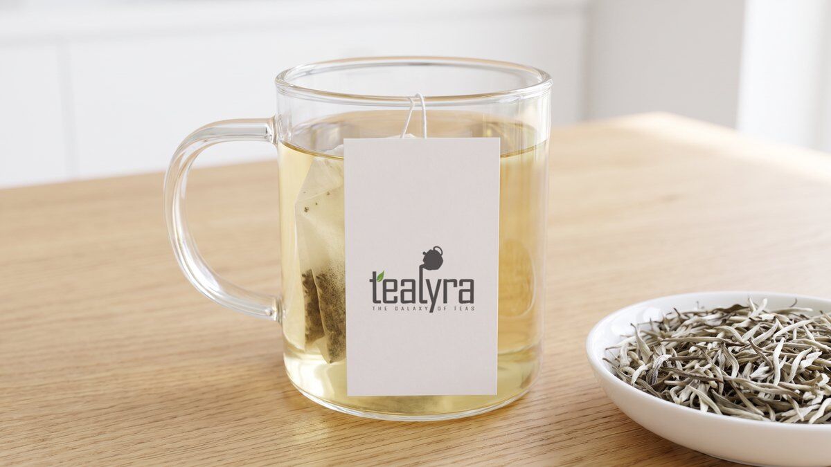 A glass mug of pale white tea with a Tealyra tea tag beside a dish of silvery Silver Needle buds
